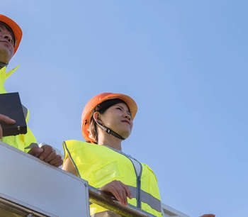 A man and woman in fluro vests and hard hats overlook a factory