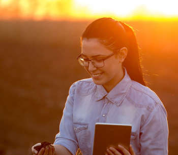 A woman stands in a field at sunset, she holds an ipad in one hand and a handful of soil in the other.