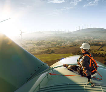 A man in a harness and hard hat sits on top of a wind turbine, overlooking a sun soaked valley