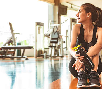 A woman in gym clothes, holding a water bottle, sits on the floor of a gym and looks off into the distance 