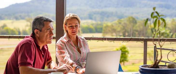A middle aged couple look at a laptop on their veranda with their farm in the background