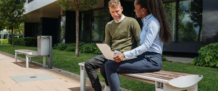 A woman and a man sit on a bench, discussing content on a shared laptop