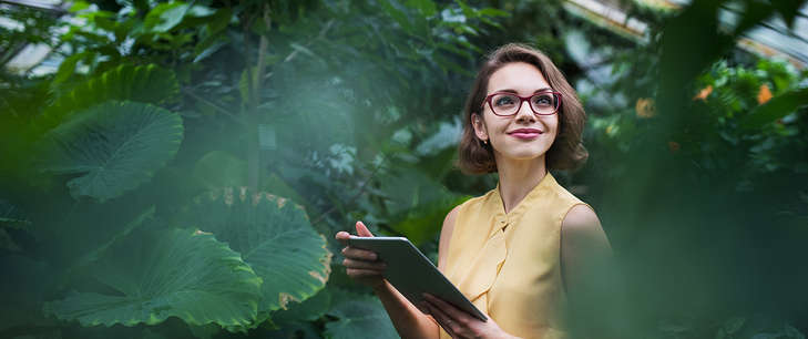 A woman, smiling and holding an iPad, stands surrounded by leaves and shrubbery in a greenhouse. 