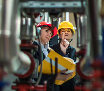 Two women in hard hats inspect machinery in a warehouse.