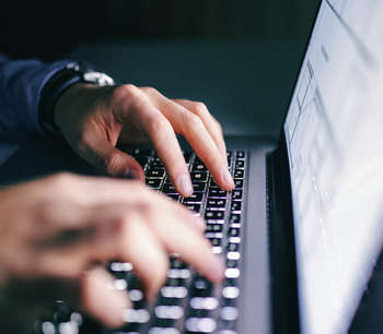 Close up of a man's hands typing on a laptop, illuminated by the laptop screen