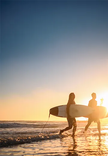 Two surfers walking on the beach with the sunset in the background.