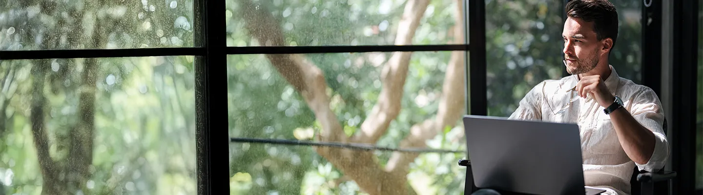 Man looking outside a window filled with green trees. 
