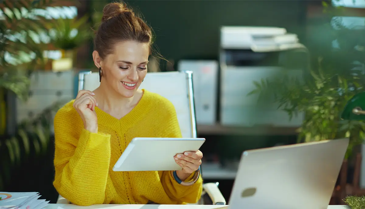 smiling modern small business owner woman in yellow sweater with tablet PC and laptop in the modern green office.