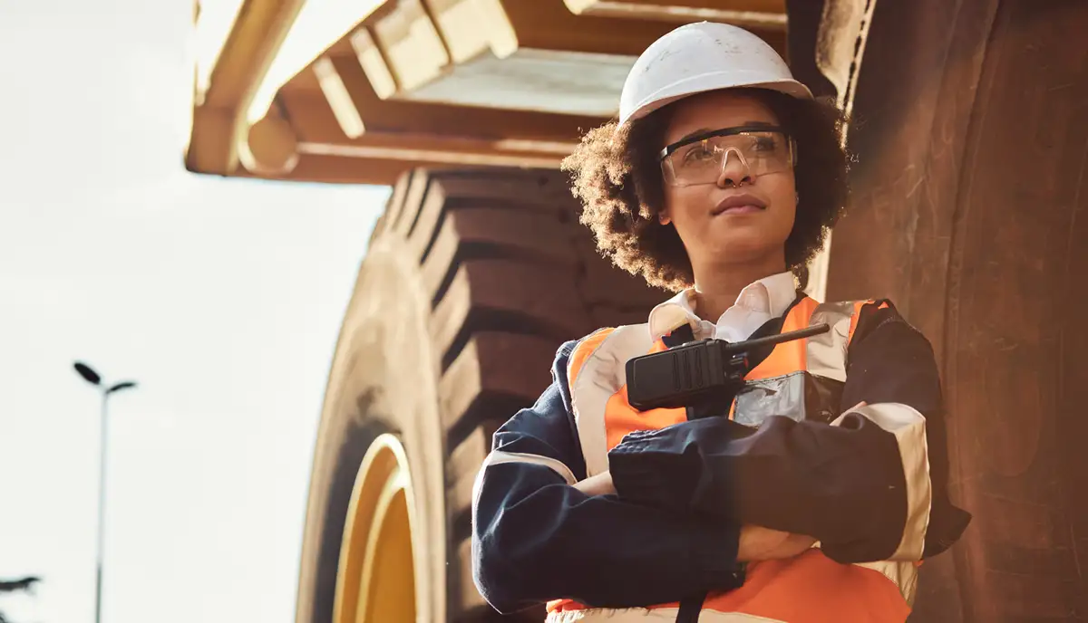 A woman in high-vis leans against a mining truck and looks off camera