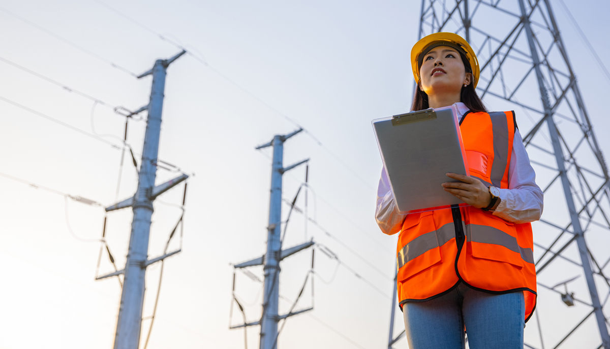 A woman in high vis stands in front of power lines and reviews a clipboard
