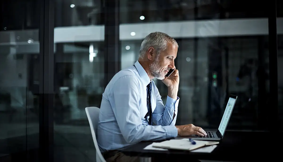 A man sits in front of his laptop computer, while talking on the phone.
