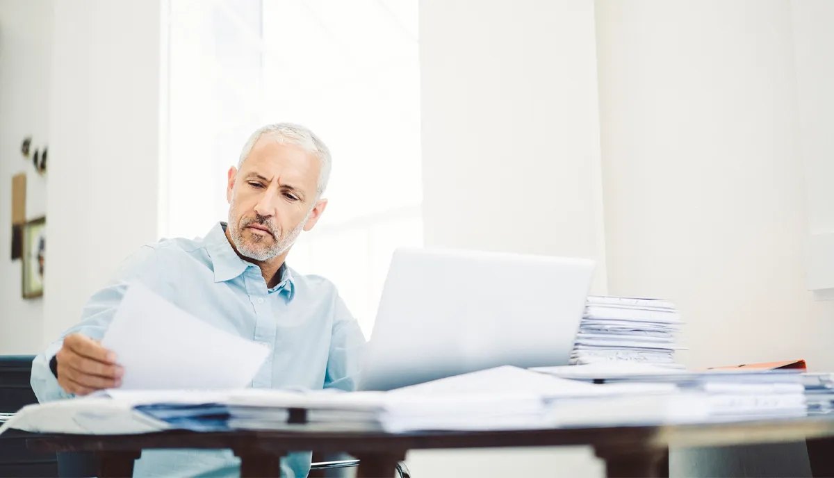 Businessman reading document at desk in office. Executive is with papers and laptop on table.