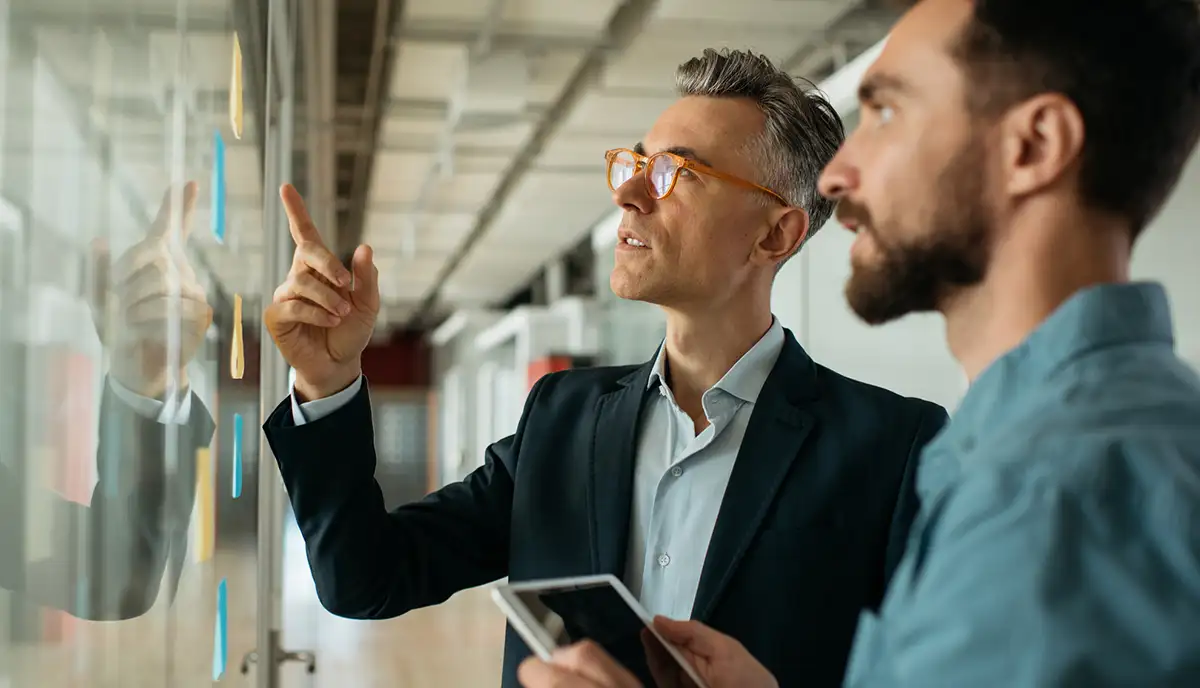 Two men, one in a suit and one in a button up, strategising on a wall map.
