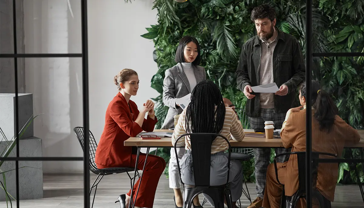 A group of people sit and stand around a table in discussion. They are in a modern workspace with a wall of greenery in the background.