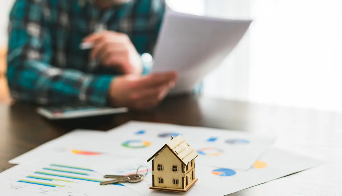 A man looking at documents pertaining to a house sale agreement. Focus on a model house on top of documents in the foreground, while the man - sitting at a table and studying the agreement papers