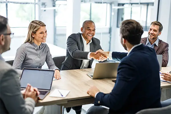 A group of business people sit around a table, two of them shake hands happily closing a deal