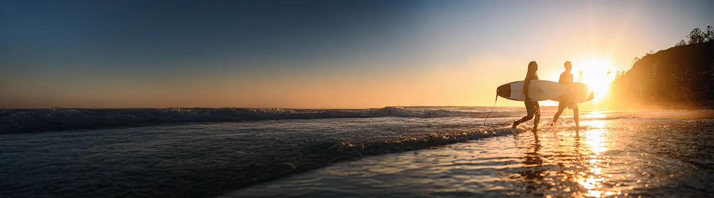 Two surfers walking on the beach with the sunset in the background.