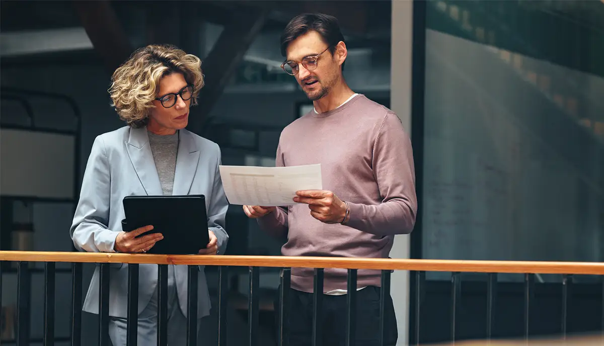 Business people reading a document together. Two professionals analyse a tax report in an office. Business colleagues discuss with each other while standing on an interior balcony.