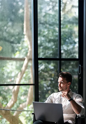 Man looking outside a window filled with green trees. 